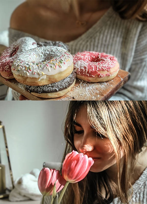 Kobieta w swetrze wącha dwa różowe tulipany; w tle deska z lukrowanymi pączkami posypanymi cukrem pudrem.