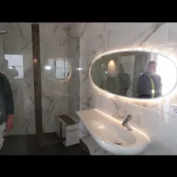 Nowoczesna łazienka z marmurowymi płytkami, podświetlanym lustrem owalnym i umywalką zintegrowaną z blatem, widoczny fragment kabiny prysznicowej z mozaiką.