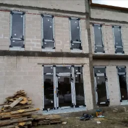 Surowy stan budynku z zamontowanymi oknami PCV i aluminiowymi drzwiami balkonowymi, zabezpieczonymi taśmą montażową, widoczne materiały budowlane na ziemi.