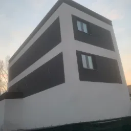 Nowoczesny budynek z elewacją w geometryczne wzory, biało-szara kolorystyka, widoczne okna, budynek w otoczeniu zieleni i innych zabudowań.