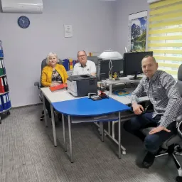 Trzy osoby w biurze, kobieta w żółtej marynarce siedzi przy biurku z segregatorami w tle, mężczyzna w okularach siedzi obok niej, a drugi mężczyzna w kraciastej koszuli siedzi z boku, uśmiechając...