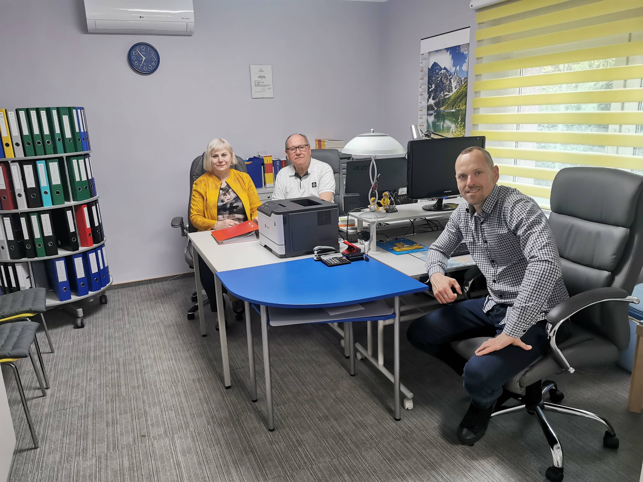 Trzy osoby w biurze, kobieta w żółtej marynarce siedzi przy biurku z segregatorami w tle, mężczyzna w okularach siedzi obok niej, a drugi mężczyzna w kraciastej koszuli siedzi z boku, uśmiechając...