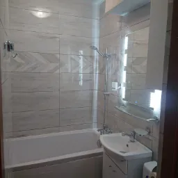 Jasna łazienka z drewnopodobnymi płytkami ułożonymi w jodełkę na ścianie, biała wanna, umywalka z szafką i podświetlane lustro z półką.