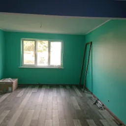 Pomalowane na turkusowo wnętrze pokoju z panelami podłogowymi, widoczne narzędzia malarskie i karton z materiałami budowlanymi, fragment ściany pomalowany na granatowo.