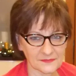 Portret kobiety w okularach i czerwonej bluzce, z małym stroikiem świątecznym w tle.