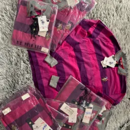 Stos ubrań marki U.S. Polo Assn. w odcieniach różu i fioletu, część zapakowana w folię, leżących na puszystym, jasnoszarym dywanie.