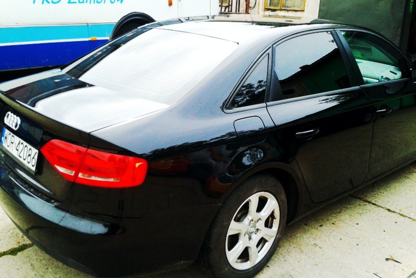 Czarne Audi A4 z przyciemnionymi tylnymi szybami, zaparkowane na betonowym podłożu. Widoczny fragment autobusu PKS w tle.