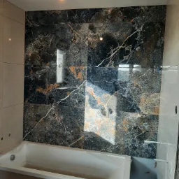 Łazienka w trakcie wykańczania: ściana z płytek imitujących czarny marmur z wyraźnym żyłowaniem w kolorze złota, biała wanna zabezpieczona folią, widoczne niedokończone fugi i wystające przewody...