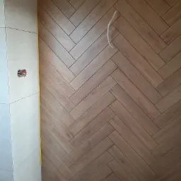 Ściana wyłożona płytkami imitującymi drewno w jodełkę, częściowo ukończona, widoczne krzyżyki dystansowe i wystający kabel, obok ściana z jasnych płytek.