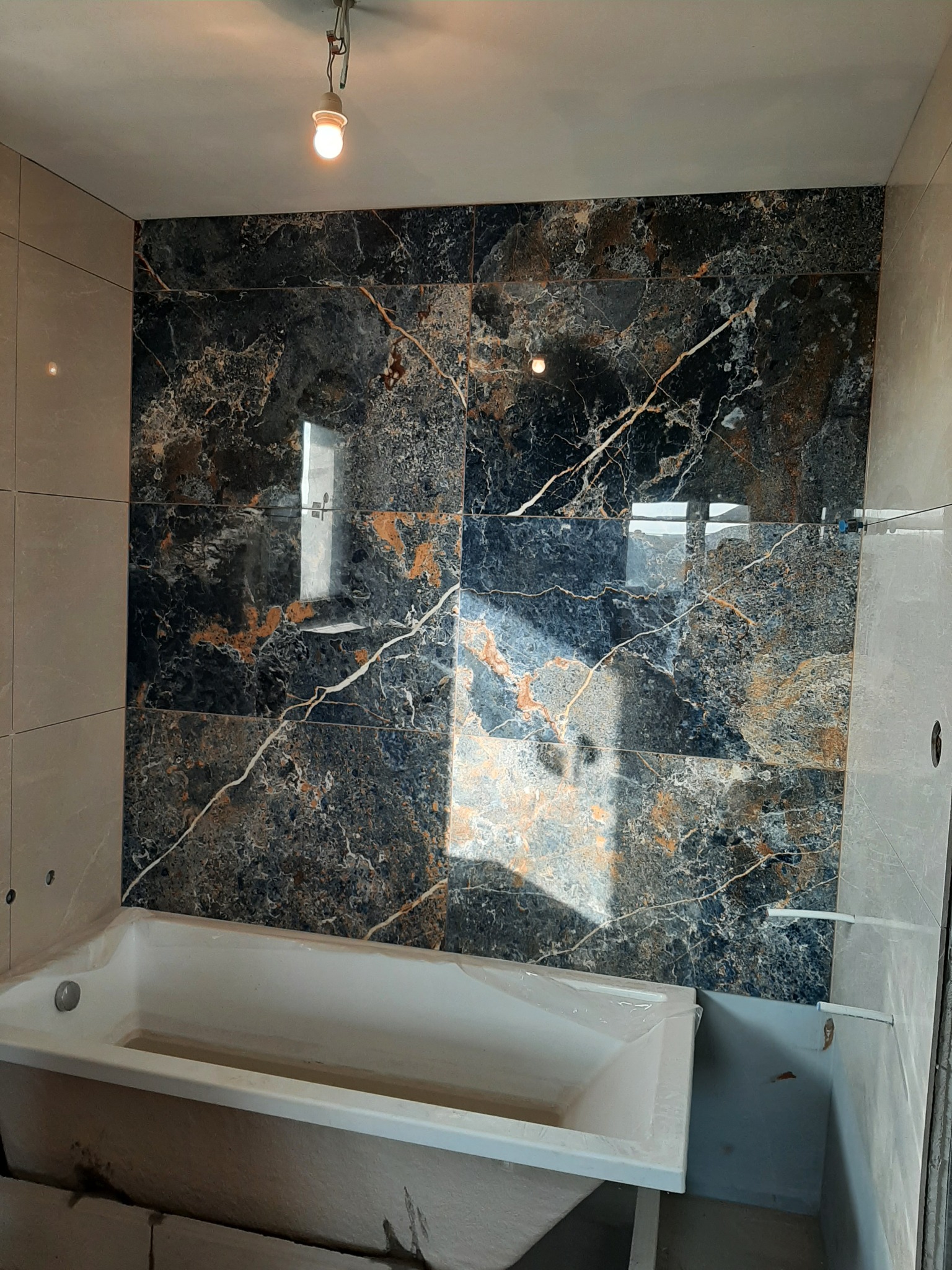 Łazienka w trakcie wykańczania: ściana z płytek imitujących czarny marmur z wyraźnym żyłowaniem w kolorze złota, biała wanna zabezpieczona folią, widoczne niedokończone fugi i wystające przewody...