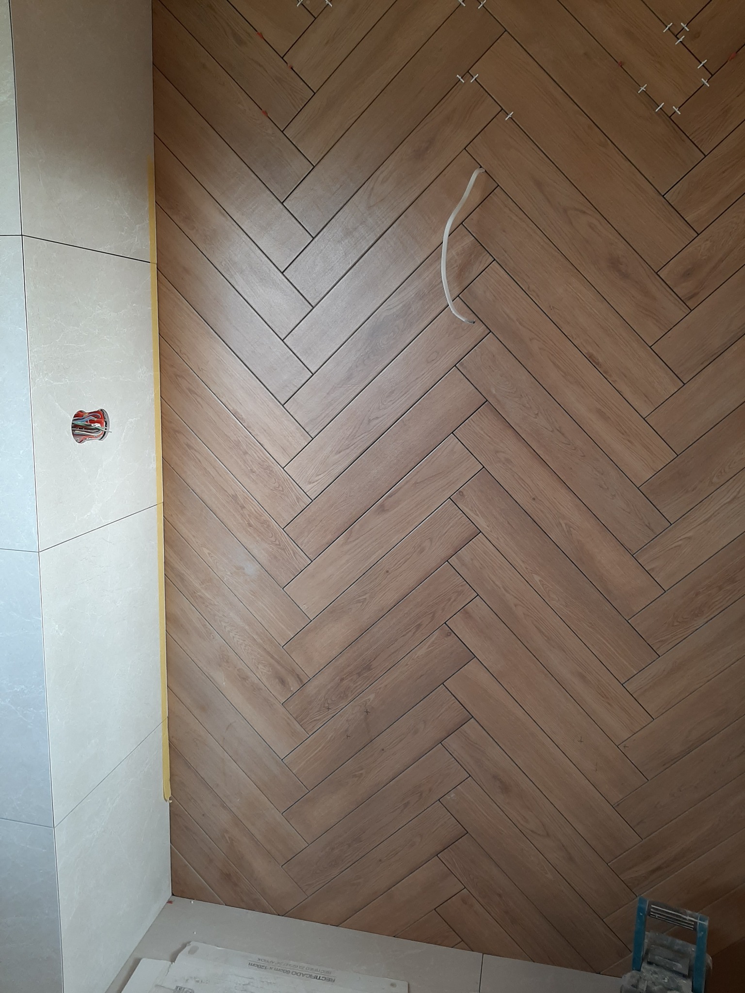 Ściana wyłożona płytkami imitującymi drewno w jodełkę, częściowo ukończona, widoczne krzyżyki dystansowe i wystający kabel, obok ściana z jasnych płytek.