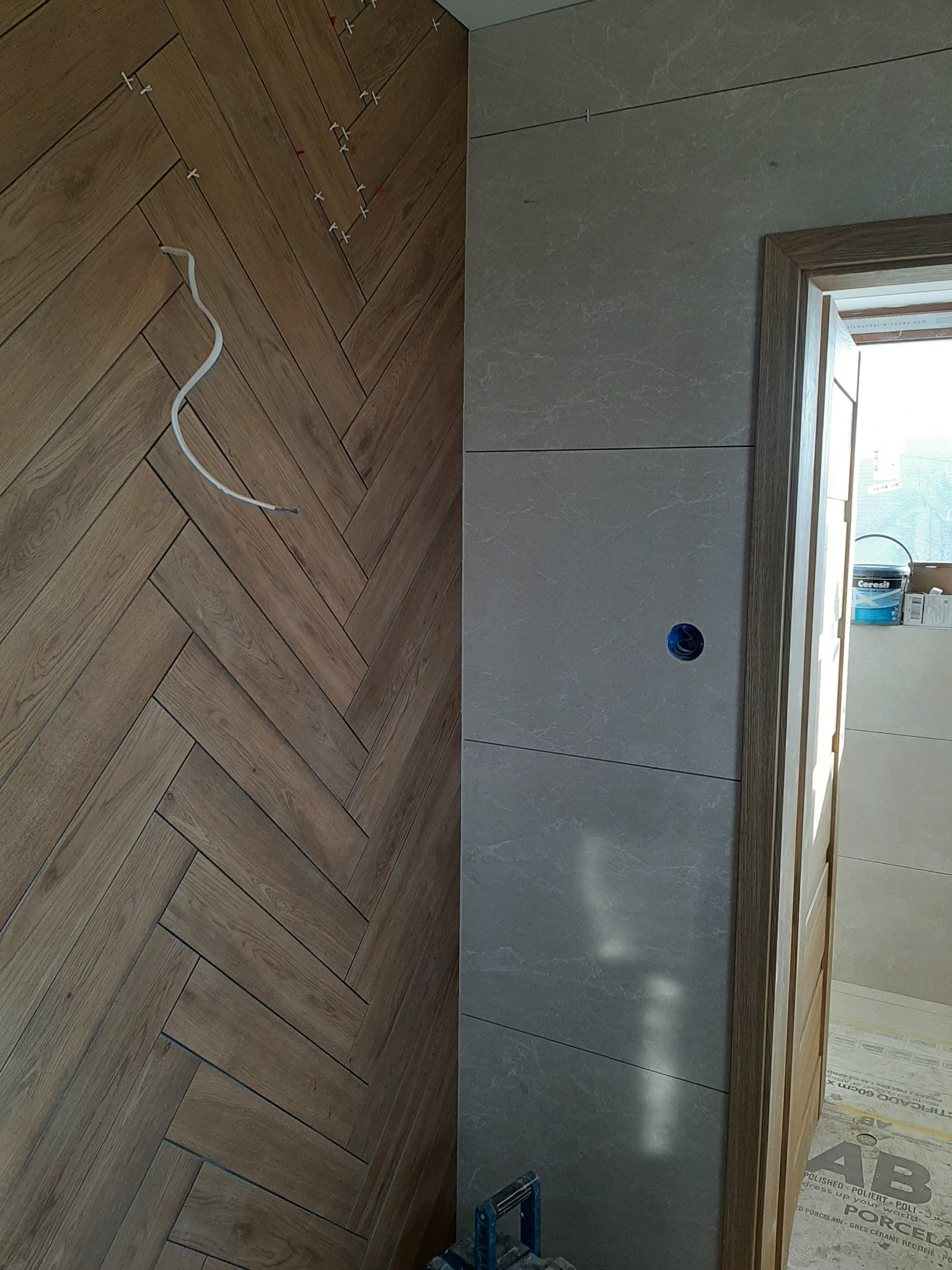 Ściana wyłożona płytkami w dwóch wzorach: drewnopodobne ułożone w jodełkę oraz szare, prostokątne płytki. Widoczne krzyżyki dystansowe, przewód elektryczny wystający ze ściany, oraz otwór...