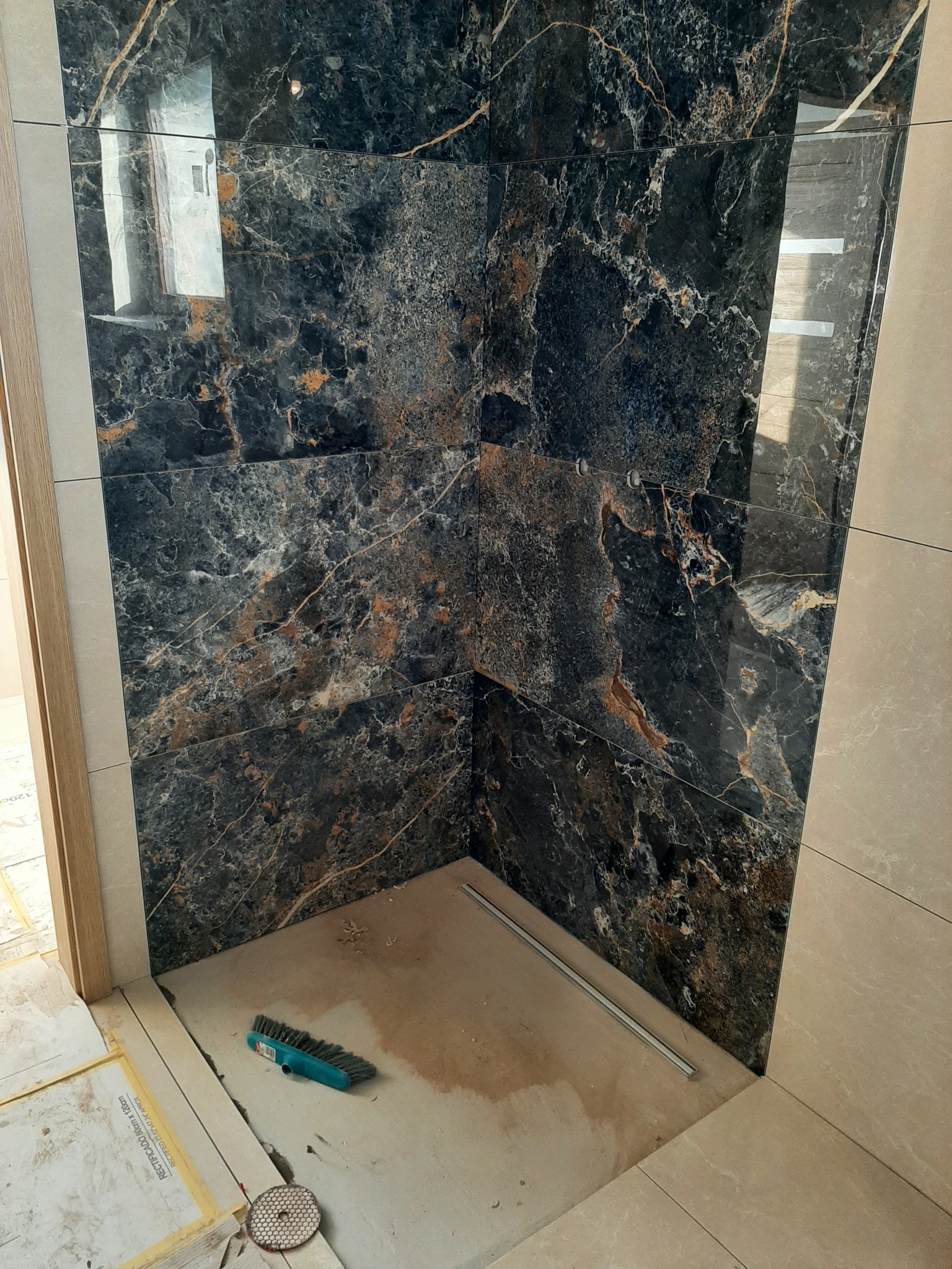 Narożnik prysznicowy wyłożony czarnymi płytkami imitującymi marmur z wyraźnymi złotymi żyłami, widoczne narzędzia glazurnicze i szczotka na podłodze, etap prac wykończeniowych.