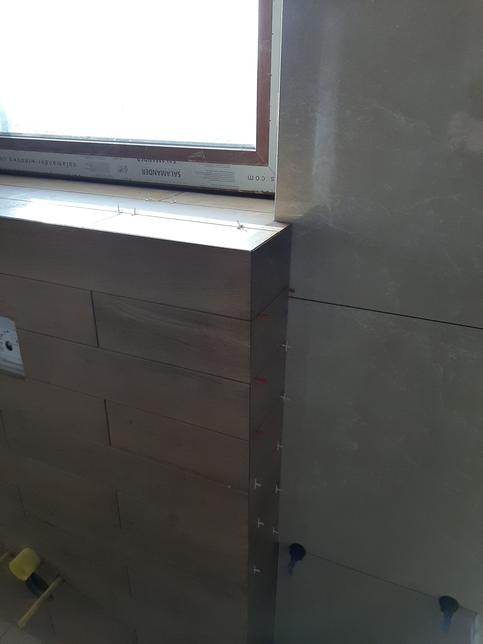 Układanie płytek imitujących drewno na ścianie w łazience, widoczne krzyżyki dystansowe i poziomica laserowa, okno z profili Salamander.