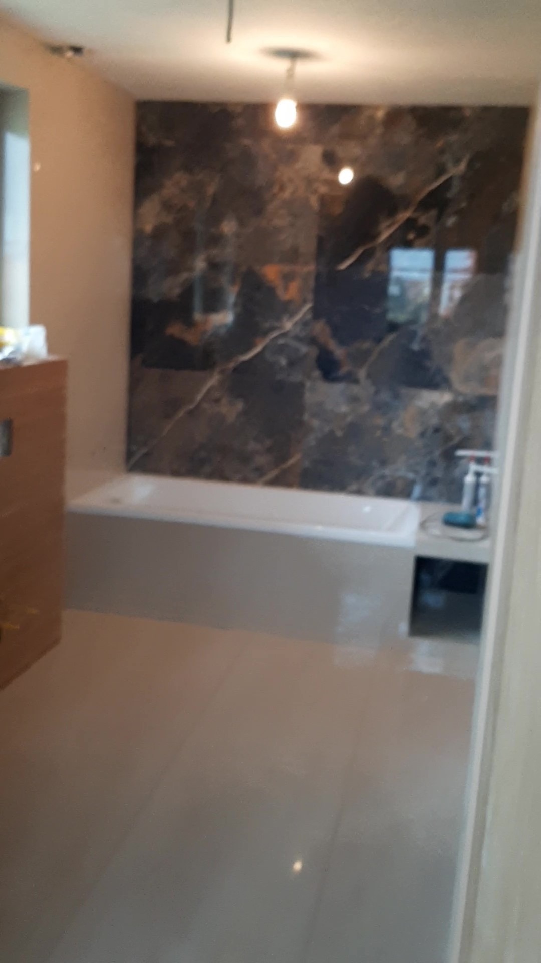 Wykończona łazienka z wanną obudowaną płytkami imitującymi marmur i beżową podłogą. Widoczne oświetlenie sufitowe i częściowo szafka.