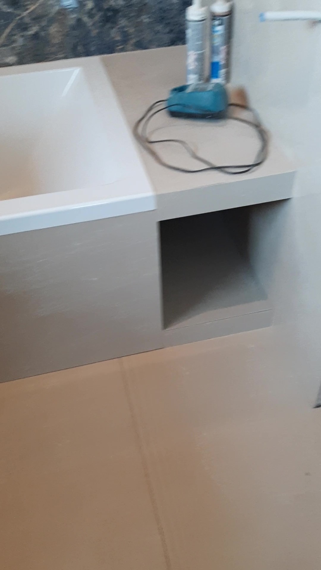 Wykończona obudowa wanny z półką i wnęką, na blacie leżą dwa pojemniki z silikonem i niebieska szlifierka.