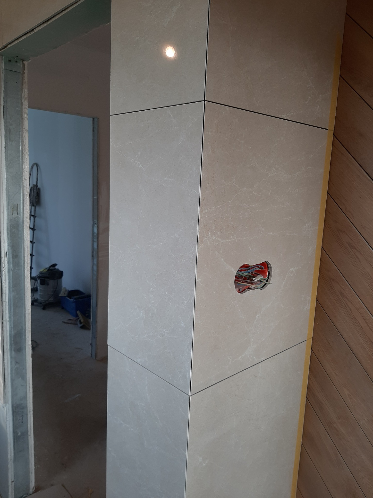 Obudowa drzwi wyłożona jasnymi płytkami imitującymi marmur, widoczne gniazdko elektryczne z przewodami, w tle widoczny odkurzacz przemysłowy i drabina oparta o ścianę, fragment parkietu ułożonego...