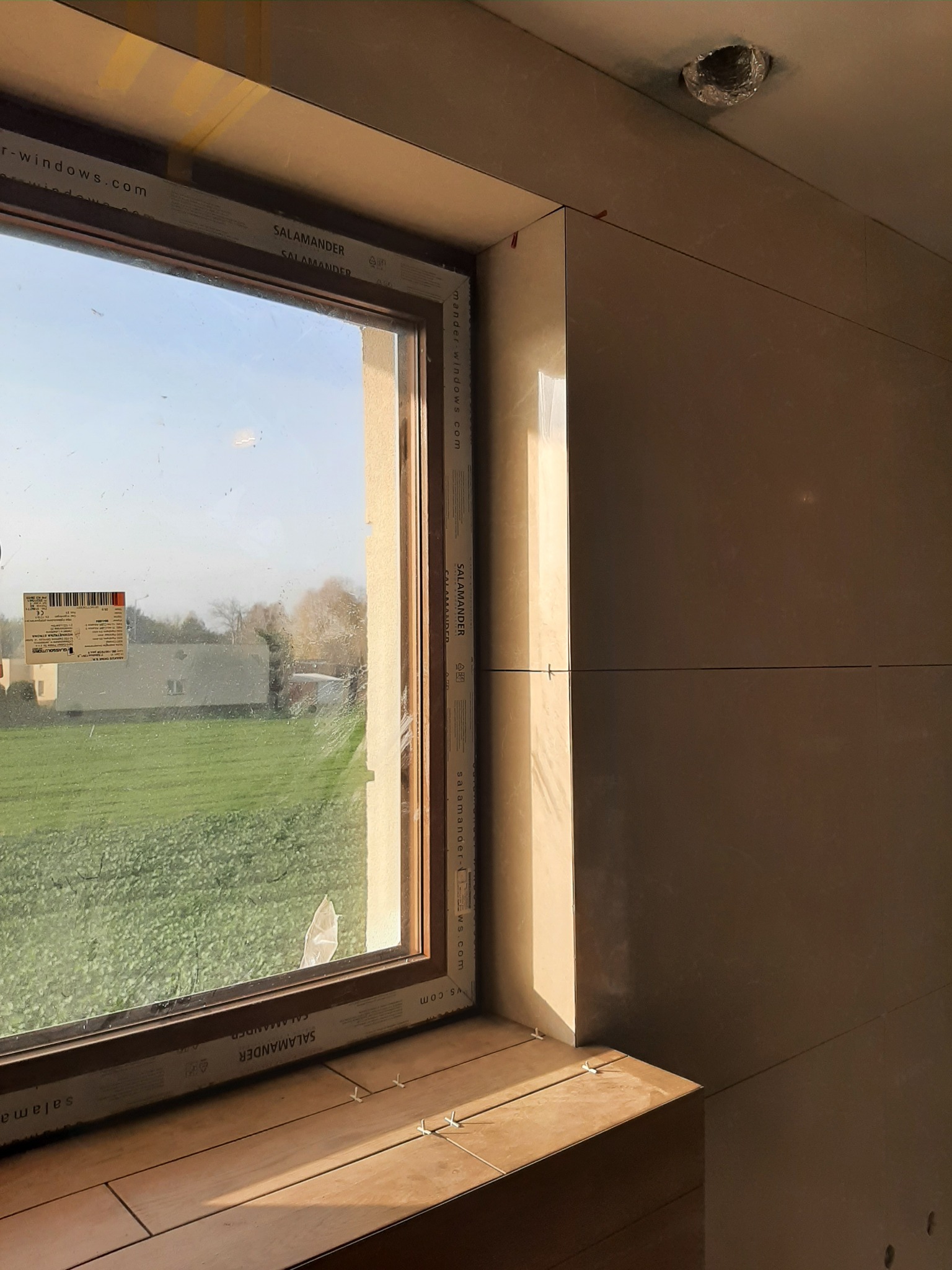 Wyłożony beżowymi płytkami parapet okienny z oknem marki Salamander, widok na zielony trawnik i dom z zewnątrz, ściana w tle wykończona płytkami, widoczne krzyżyki dystansowe między płytkami.