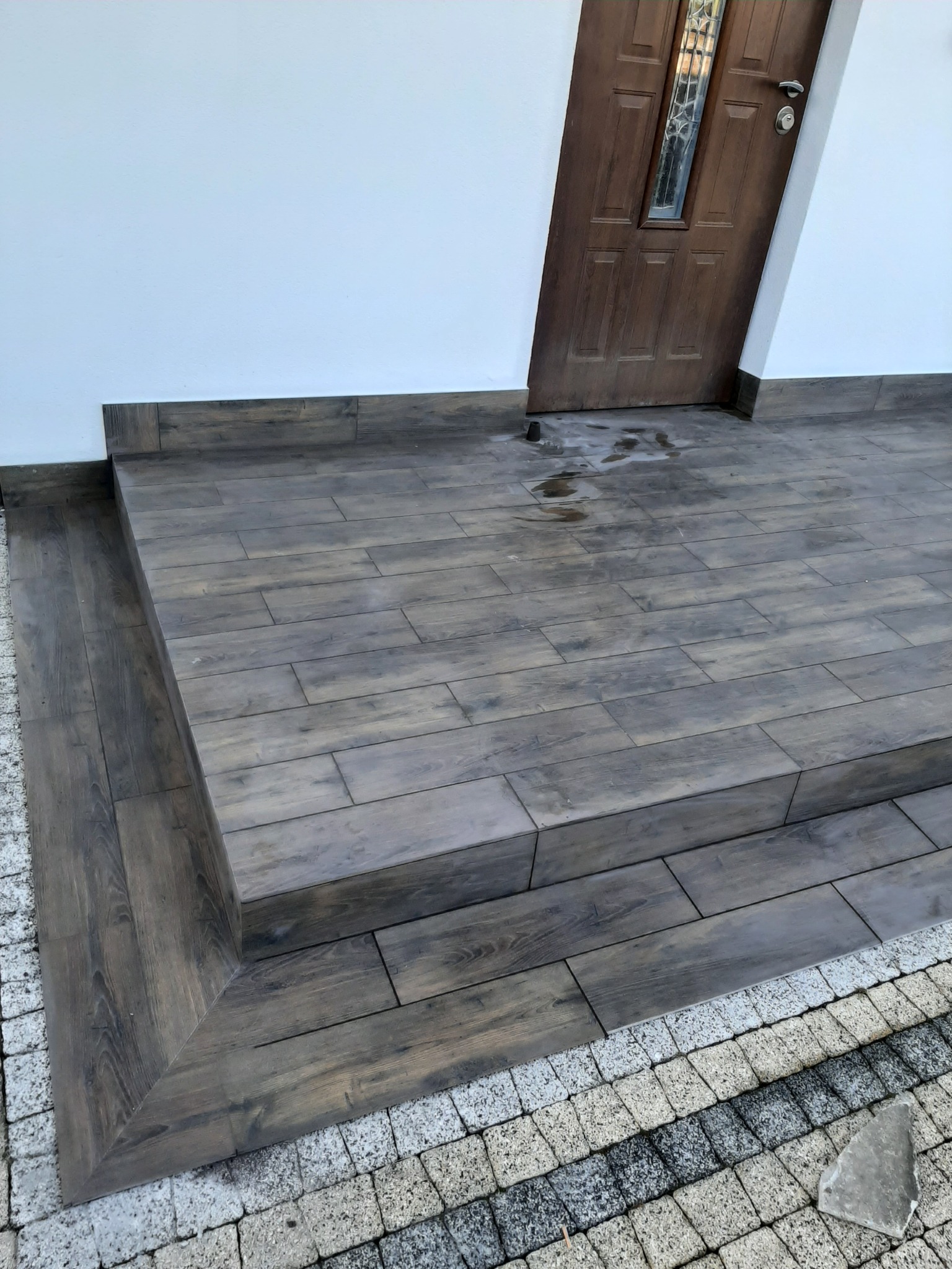 Wykończone płytkami imitującymi drewno schody wejściowe do domu z białymi ścianami i drewnianymi drzwiami, obok ułożona kostka brukowa.