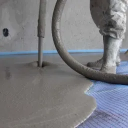 Aplikacja wylewki samopoziomującej na siatce podłogowej, widoczne gumowe buty pracownika i wąż doprowadzający mieszankę.