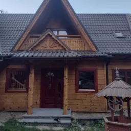 Drewniany dom w stylu góralskim z ciemnoczerwonymi ramami okiennymi i drzwiami, widoczna studnia z drewnianym kołem po prawej stronie, buty na schodach wejściowych.