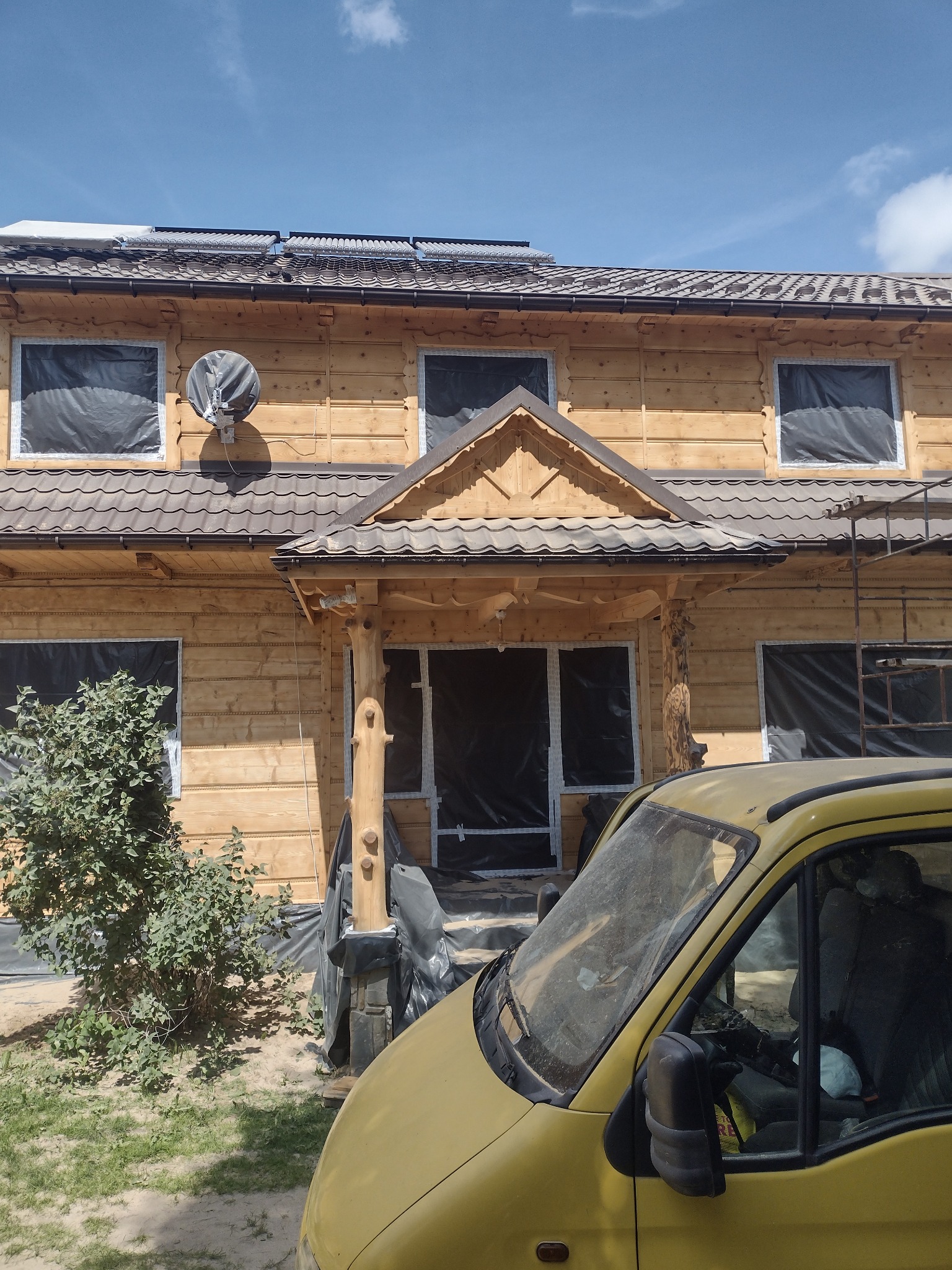 Drewniany dom z zasłoniętymi folią oknami, przygotowany do prac malarskich, z widocznym fragmentem żółtego samochodu na pierwszym planie.