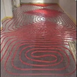 Czerwone rury ogrzewania podłogowego ułożone na folii w surowym pomieszczeniu przed wylewką, widoczne okna i ściany w stanie deweloperskim.