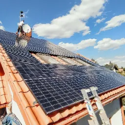 Montaż paneli fotowoltaicznych na dachu domu z pomarańczową dachówką, widoczny monter na kominie i drabina oparta o dach.