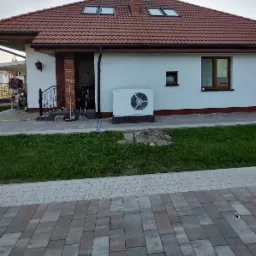 Biała pompa ciepła Toshiba zamontowana na zewnątrz białego domu z czerwoną dachówką, widoczna sterta ziemi na trawniku.