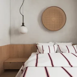 Minimalistyczna sypialnia z łóżkiem w białą pościel w paski, drewnianą szafką nocną wbudowaną w zagłówek i okrągłą, plecioną dekoracją ścienną, oświetlona wiszącą lampą z okrągłym kloszem.