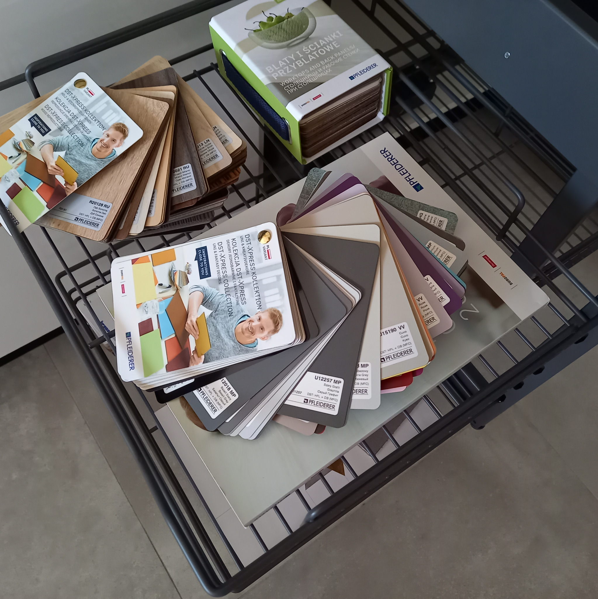 Próbniki kolorów i faktur płyt meblowych Pfleiderer, ułożone w metalowym koszu wysuwanym, prezentujące szeroką gamę odcieni drewna, szarości i beżu, z widocznymi oznaczeniami producenta i nazwami...