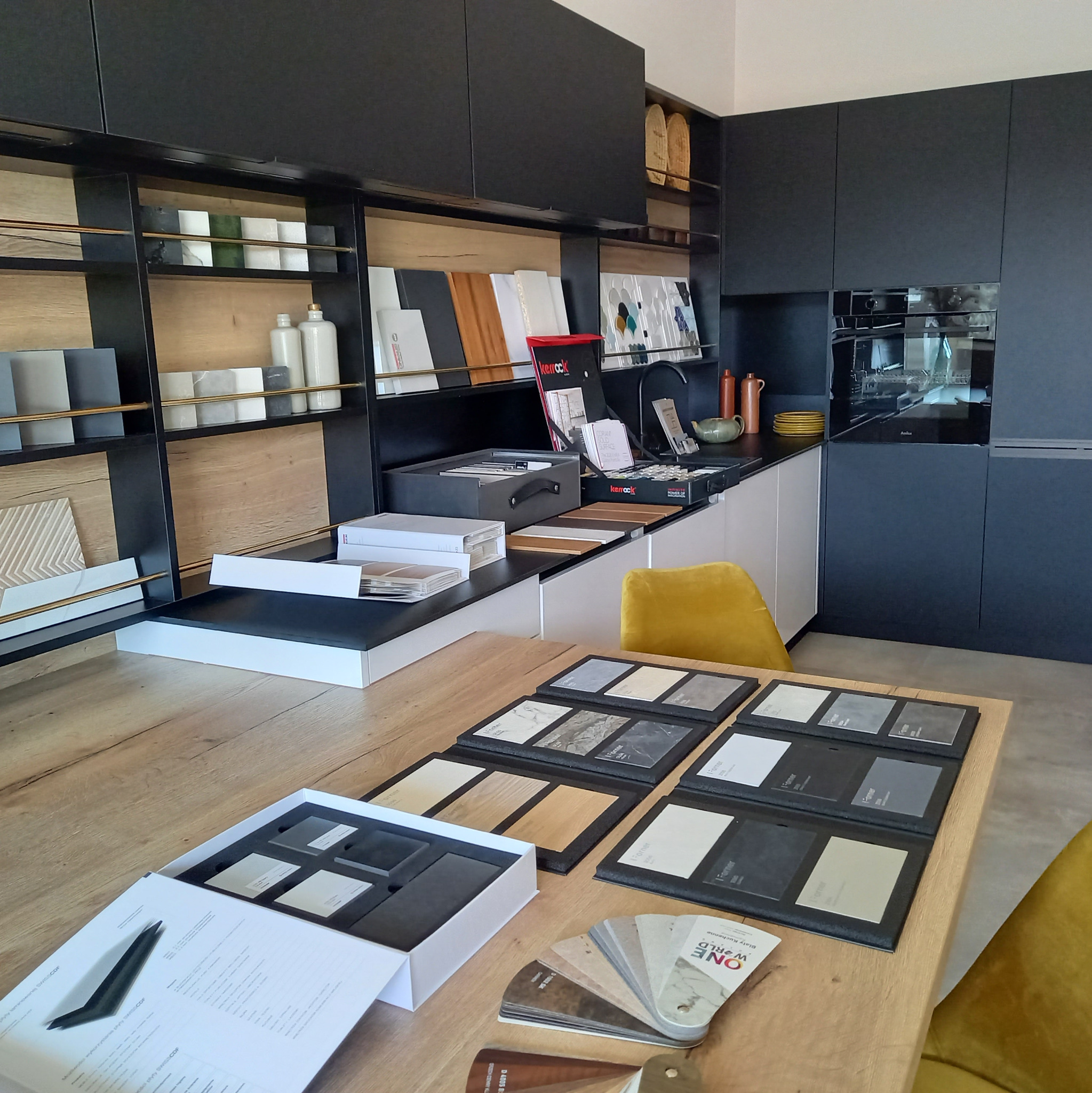 Ekspozycja próbek materiałów wykończeniowych do kuchni w showroomie: forniry, płyty laminowane, blaty, z czarnymi szafkami w tle i żółtym krzesłem.