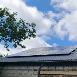 Panele fotowoltaiczne na szarym blaszanym dachu budynku z cegły, na tle błękitnego nieba z chmurami i drzewa. Widok z dołu.