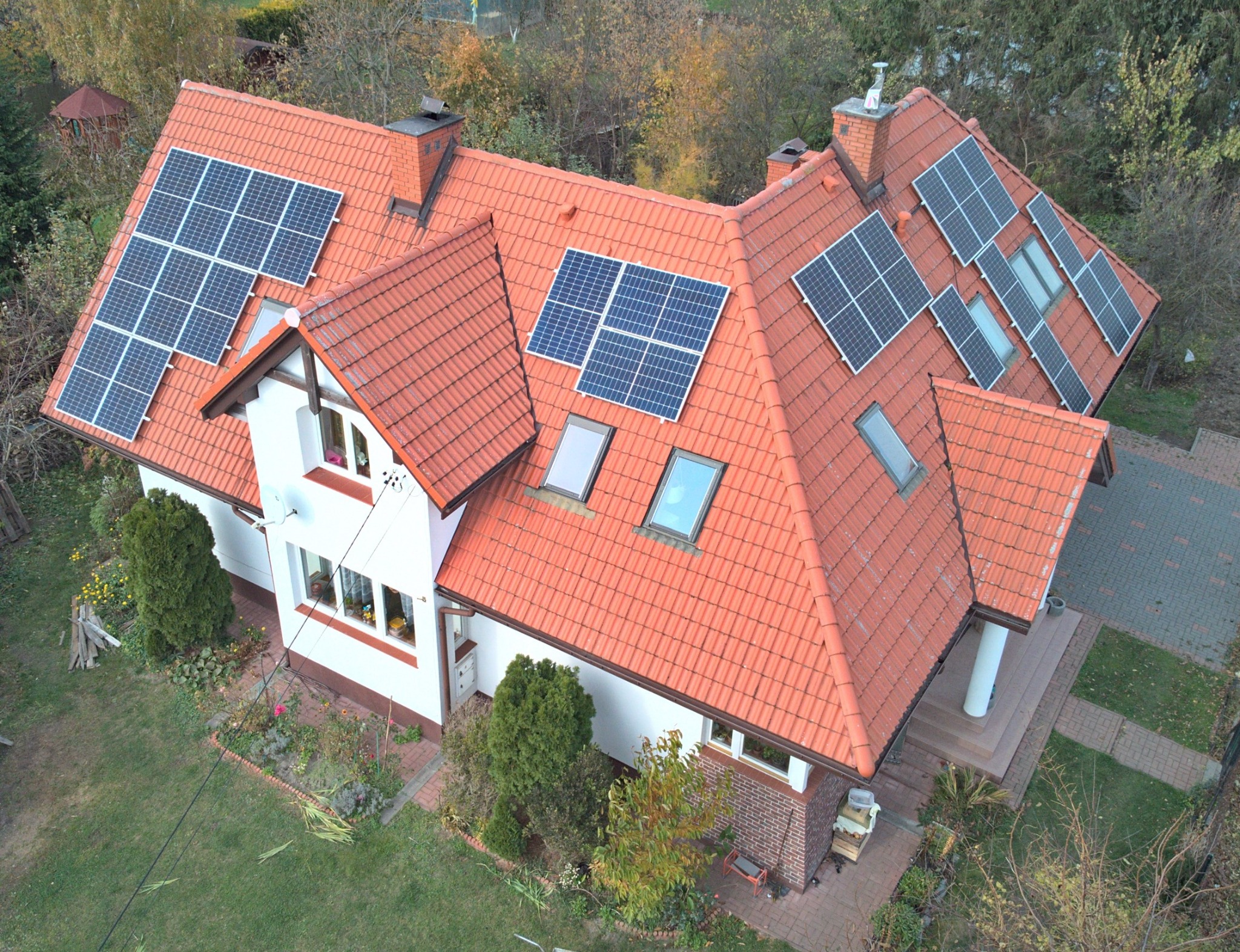 Widok z góry na dom jednorodzinny z czerwoną dachówką, na której zamontowane są panele fotowoltaiczne w różnych konfiguracjach i rozmiarach, w tle drzewa i trawnik.