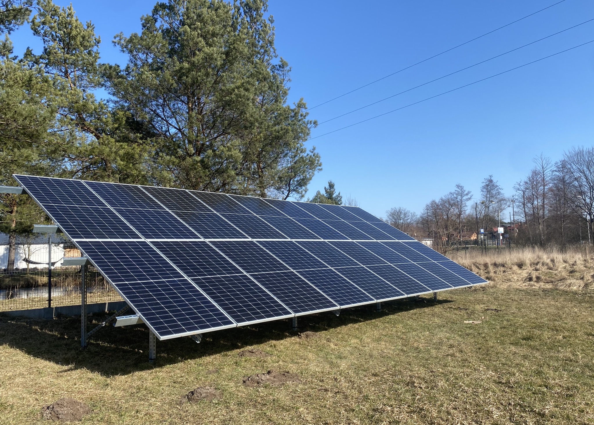 Panel fotowoltaiczny zamontowany na metalowej konstrukcji na trawiastym terenie, widoczne drzewa w tle pod błękitnym niebem.