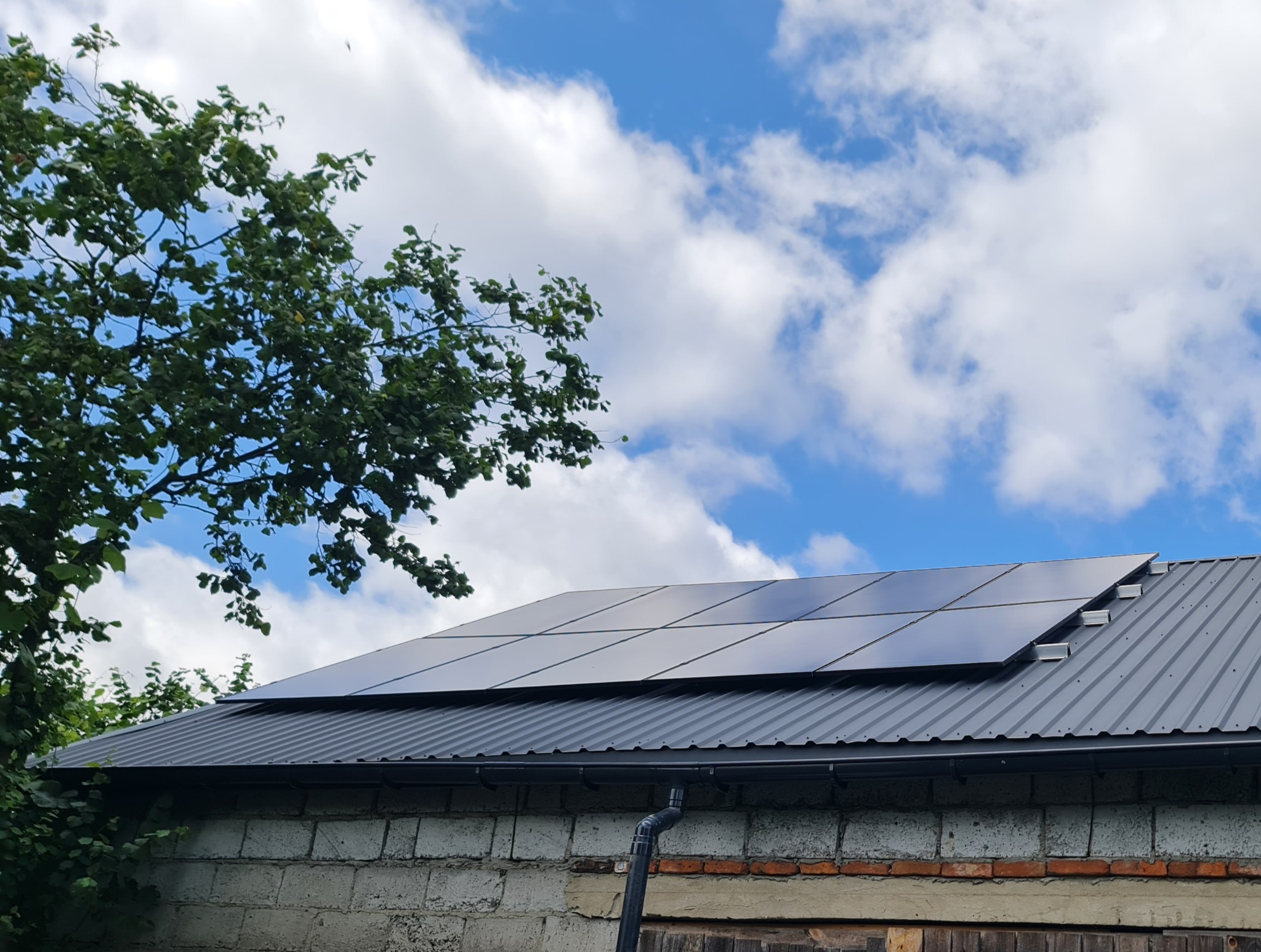 Panele fotowoltaiczne na szarym blaszanym dachu budynku z cegły, na tle błękitnego nieba z chmurami i drzewa. Widok z dołu.