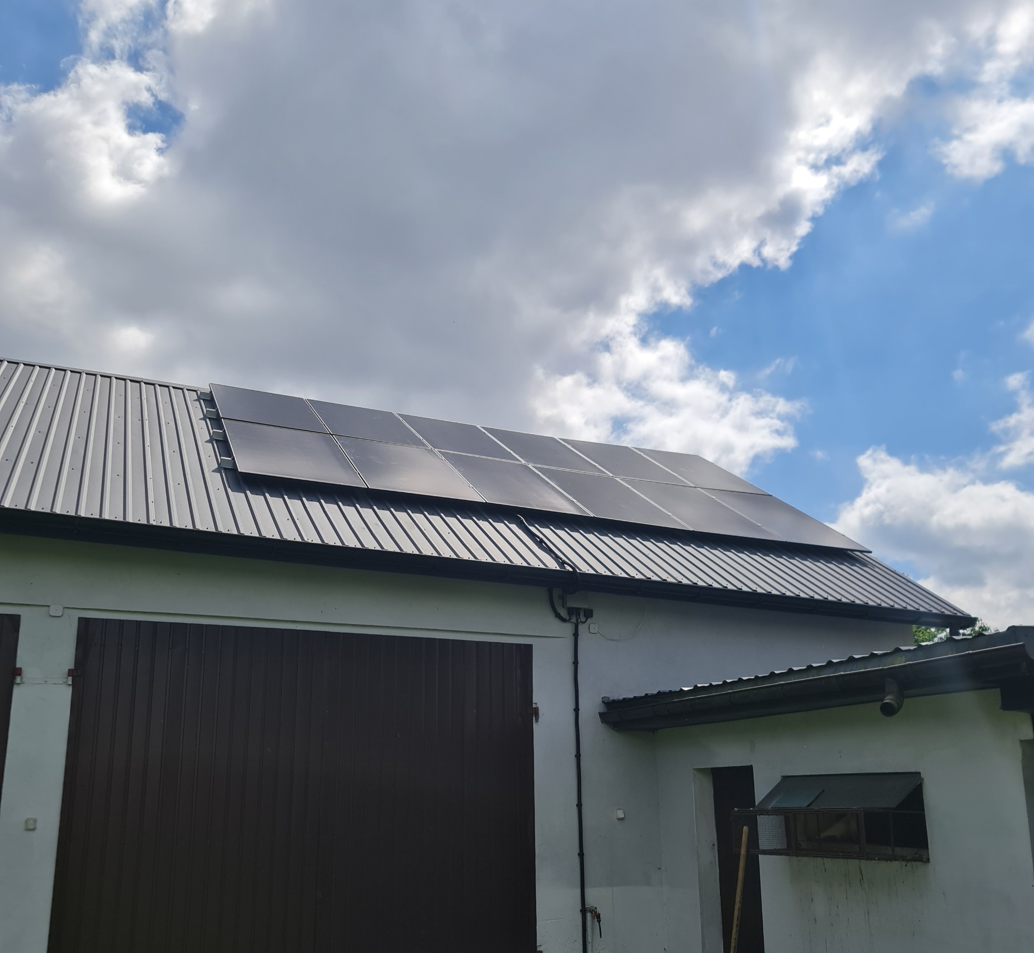 Czarny panel fotowoltaiczny na metalowym dachu budynku gospodarczego z brązową bramą garażową, na tle błękitnego nieba z chmurami.