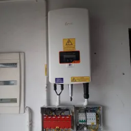 Zamontowany inwerter fotowoltaiczny Solis na białej ścianie, z widocznymi zabezpieczeniami w otwartych puszkach i przewodami. Dodatkowo widoczna skrzynka elektryczna po lewej stronie.