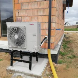 Zewnętrzna jednostka Bosch na czarnym metalowym stelażu, zamontowana przy ścianie budynku w trakcie budowy, widoczne cegły, izolacja fundamentów oraz żółty peszel.