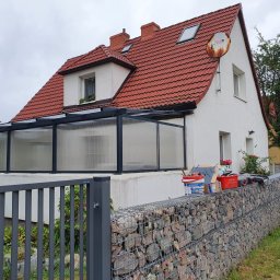 PPUH Aplauz - Dom z czerwonym dachem i przeszklonym tarasem, częściowo zasłonięty gabionowym ogrodzeniem z narzędziami i farbami na wierzchu.