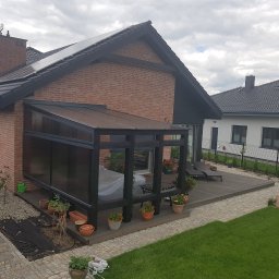 PPUH Aplauz - Zadaszony taras z czarną konstrukcją przylegający do ceglanego domu, z drewnianym podestem, kwiatami w donicach i leżakami, widok na ogród i sąsiedni dom z szarą dachówką.