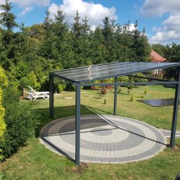 PPUH Aplauz - Metalowa pergola z przezroczystym zadaszeniem na okrągłym, wybrukowanym patio w ogrodzie, w tle drzewa iglaste i leżaki.