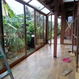 Zadaszony taras z rozsuwanymi szklanymi drzwiami, widok na bujny ogród, drewniane słupy konstrukcyjne, częściowo wykończona podłoga z płytek imitujących drewno, drabina oparta o ścianę i narzędzia...