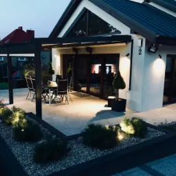 Oświetlony taras z pergolą przy domu z ciemną blachą trapezową na dachu, widoczne rośliny ozdobne w donicach i rabatach.