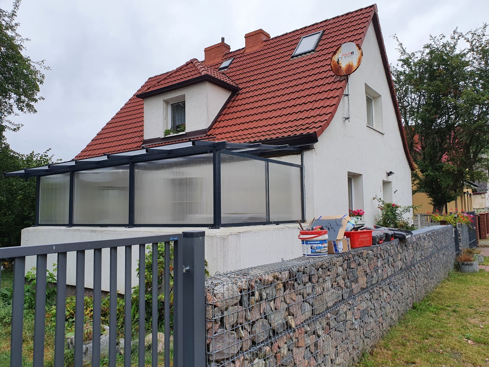 Dom z czerwonym dachem i przeszklonym tarasem, częściowo zasłonięty gabionowym ogrodzeniem z narzędziami i farbami na wierzchu.