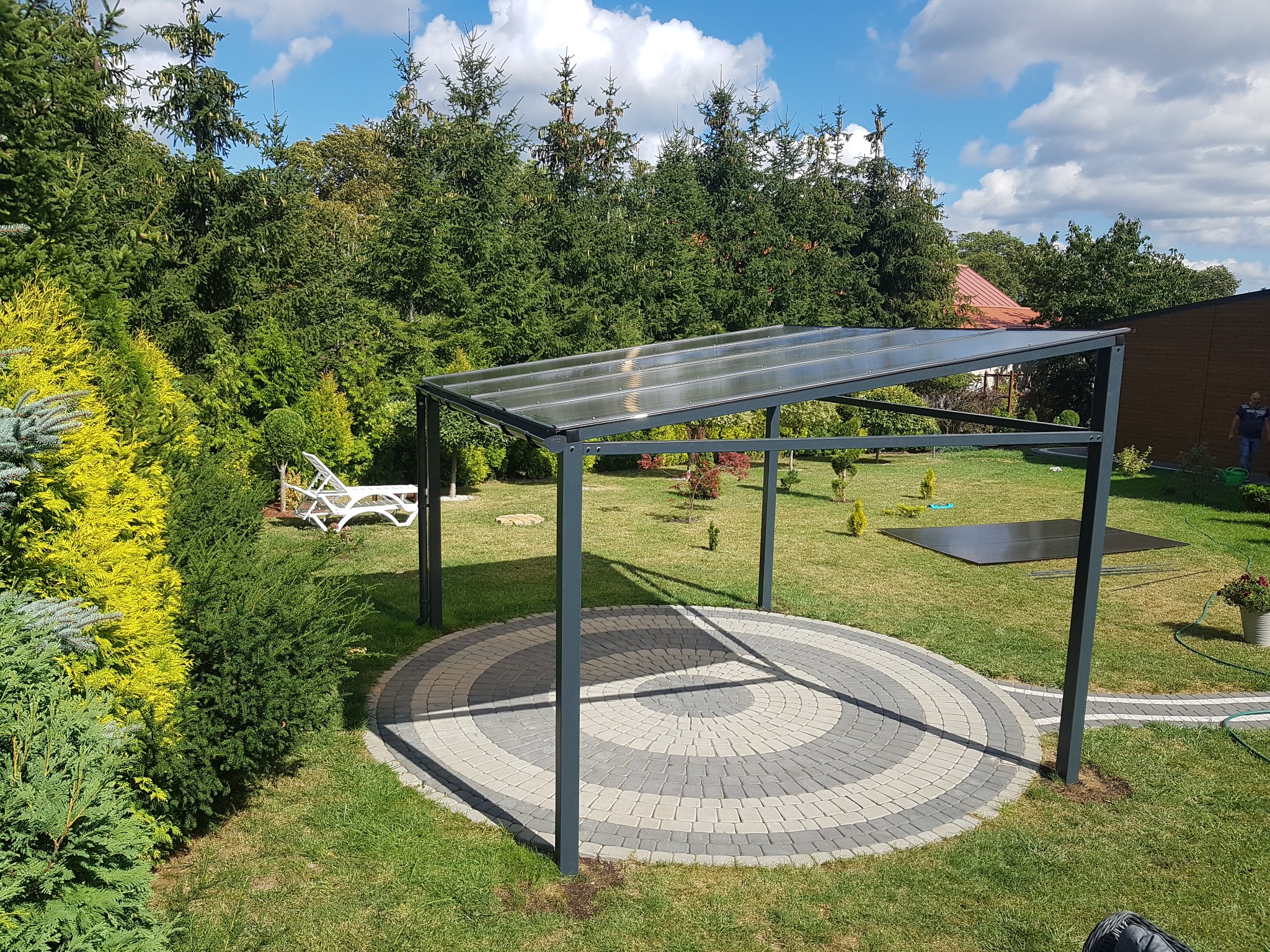 Metalowa pergola z przezroczystym zadaszeniem na okrągłym, wybrukowanym patio w ogrodzie, w tle drzewa iglaste i leżaki.