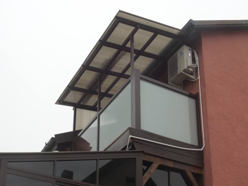 Zadaszony balkon z przeszkloną balustradą, brązowa konstrukcja drewniana, widoczny klimatyzator na ścianie budynku.