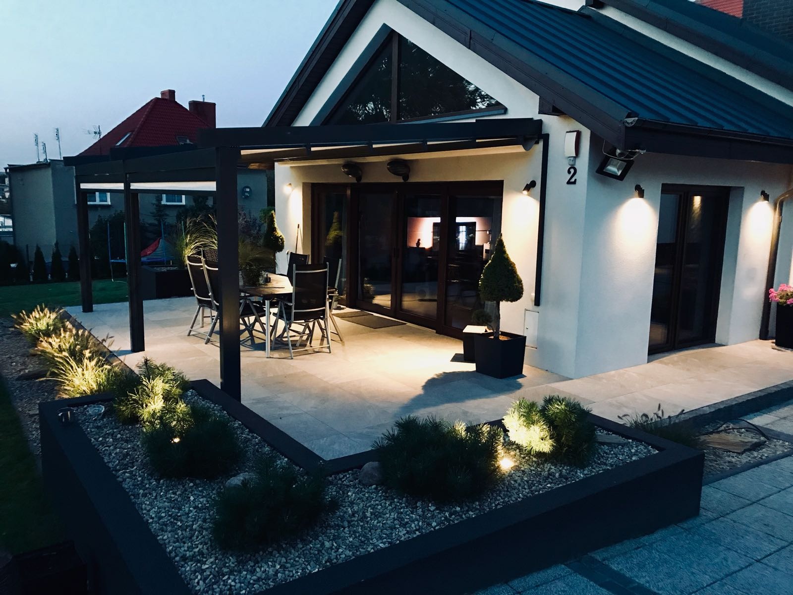 Oświetlony taras z pergolą przy domu z ciemną blachą trapezową na dachu, widoczne rośliny ozdobne w donicach i rabatach.
