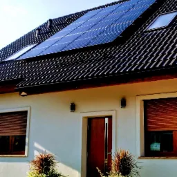 Panele słoneczne na dachu domu z ciemną dachówką, widoczne okna dachowe i rolety zewnętrzne.