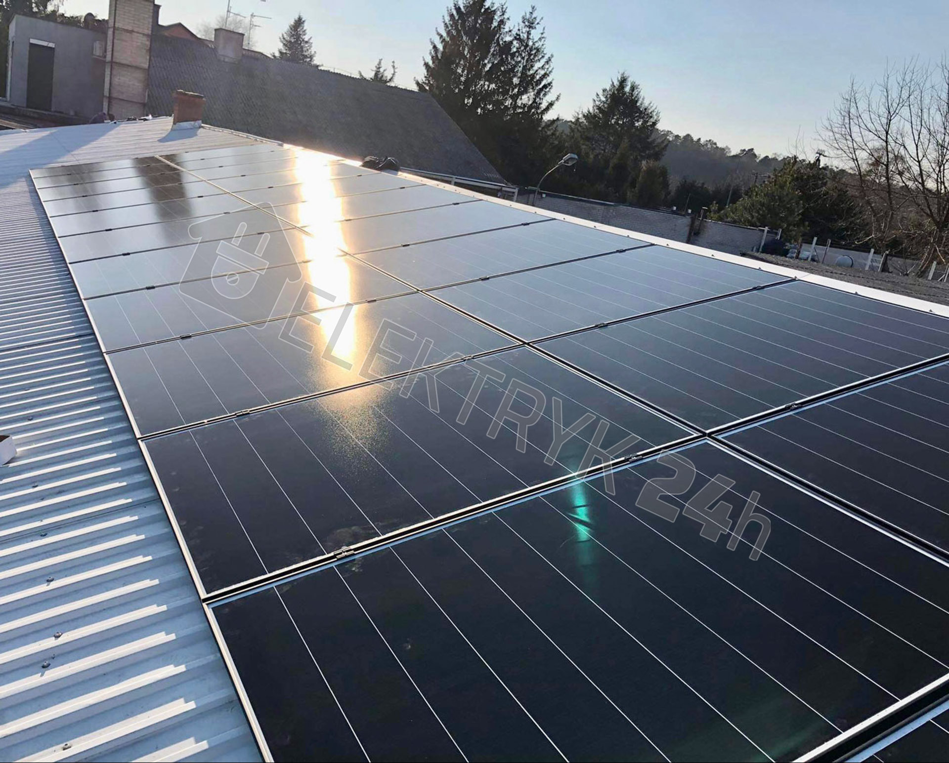 Panele fotowoltaiczne zamontowane na blaszanym dachu domu jednorodzinnego w słoneczny dzień, widoczne odbicia światła na powierzchni paneli.